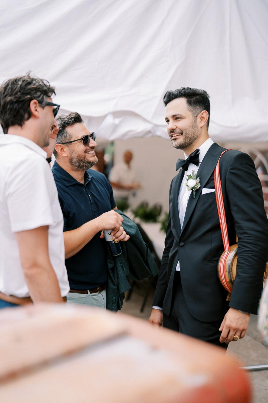 A groom in a tuxedo with a boutonniere talks and smiles with two male wedding guests under a tent.