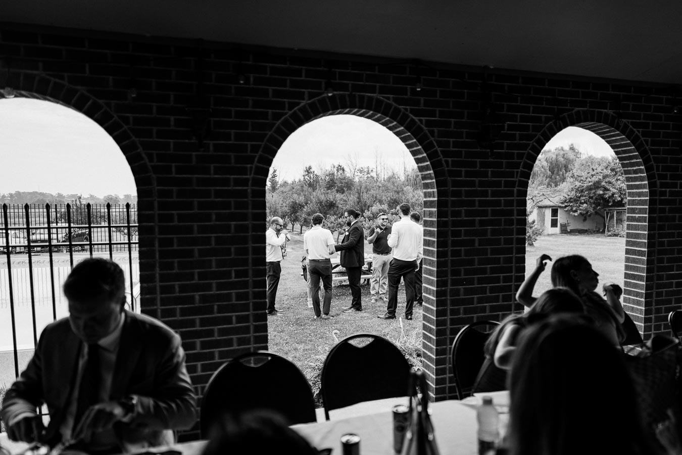Black and white image of a social gathering, viewed through brick archways. People in formal attire are engaged in conversation on a lawn outside, surrounded by trees and a small building. Indoor seating and a table are visible in the foreground.