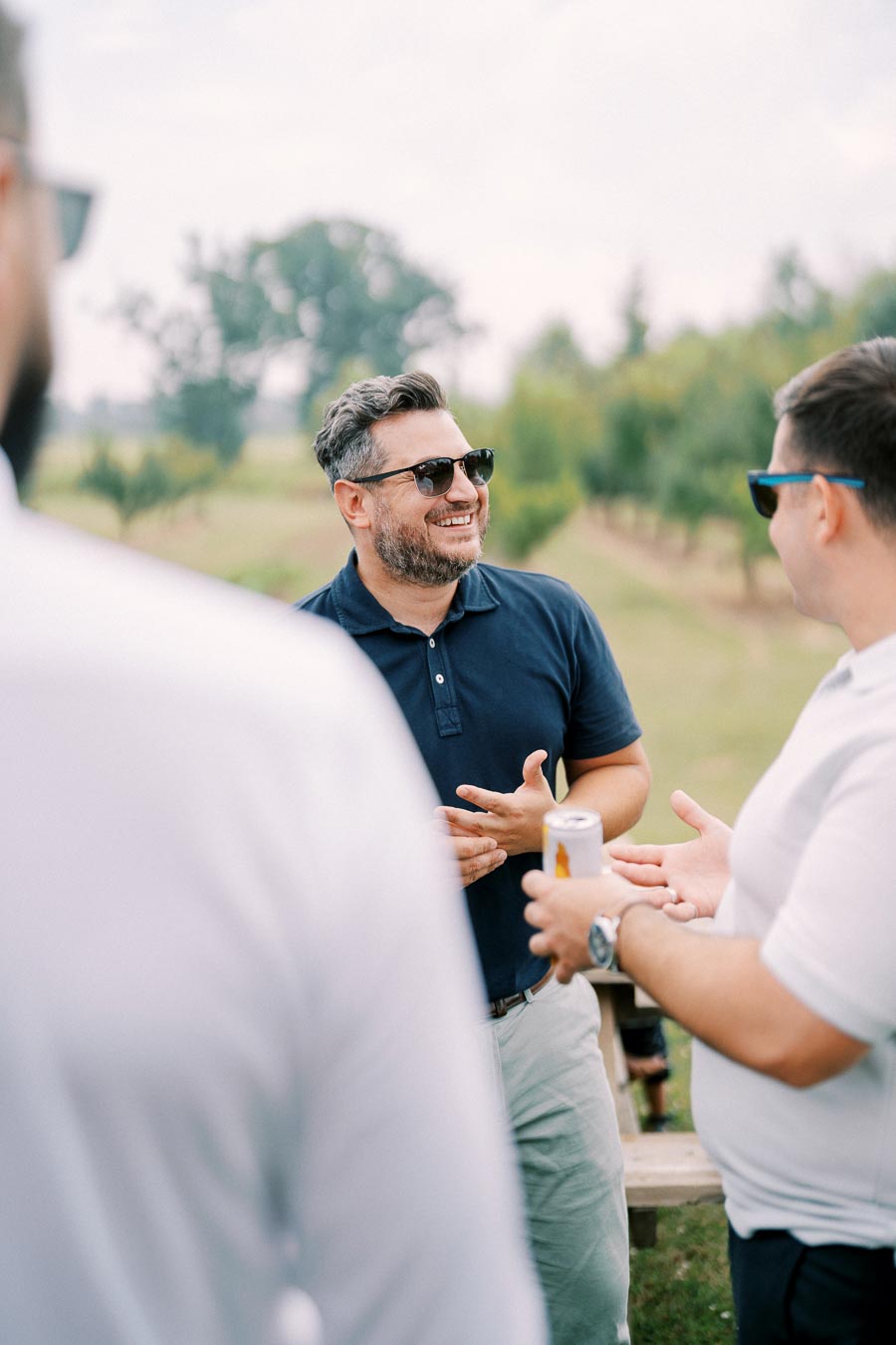 Men enjoying a conversation outdoors while holding drinks, wearing sunglasses, with trees in the background.
