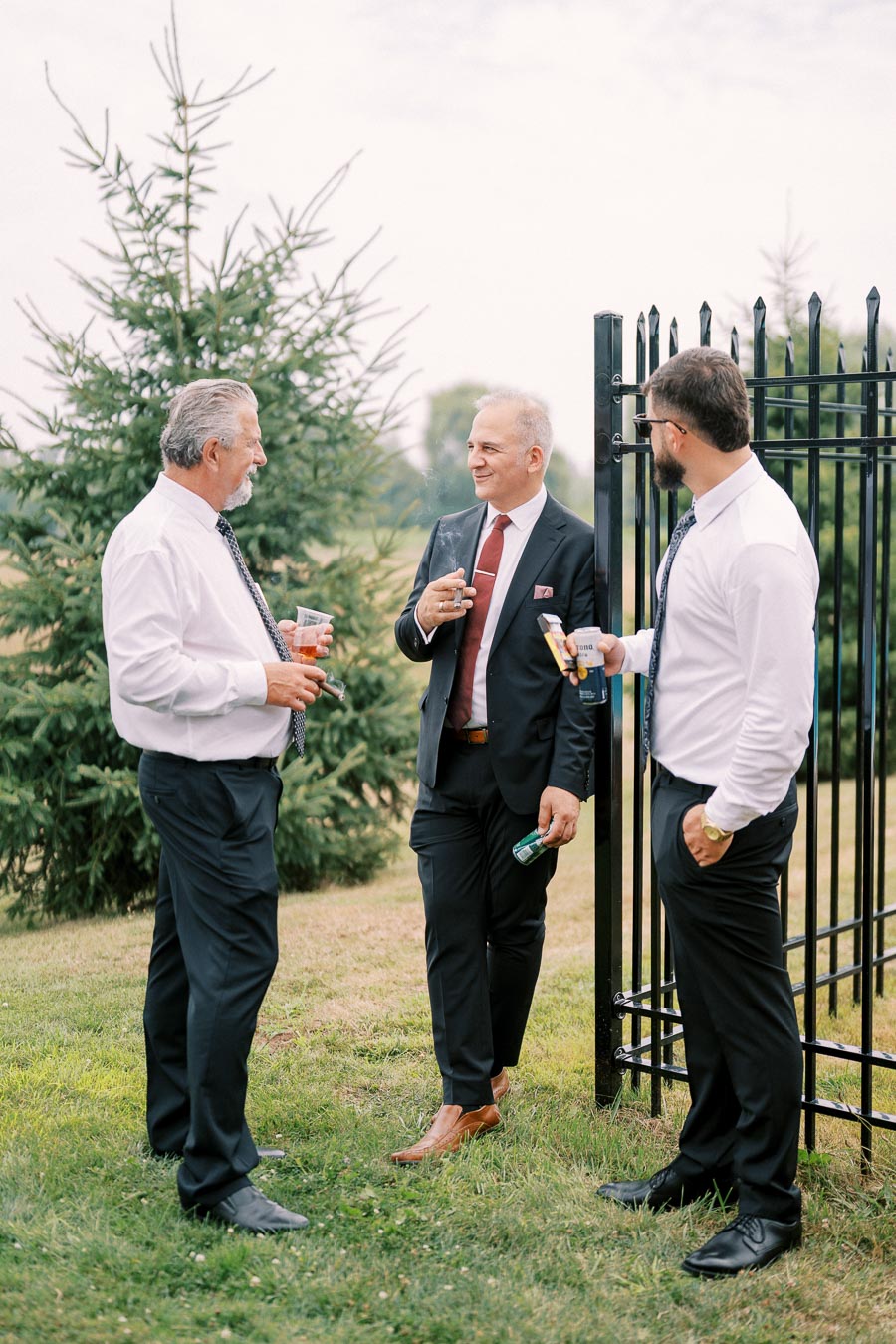 Three men in formal attire enjoying a conversation outdoors, holding drinks and cigars, near a black metal fence and evergreen trees.