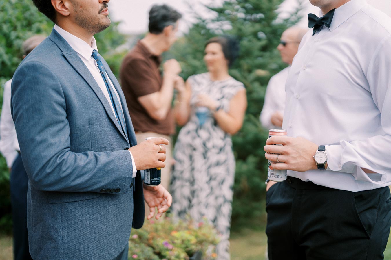 Men in formal attire holding cans at an outdoor social gathering with people chatting in the background.