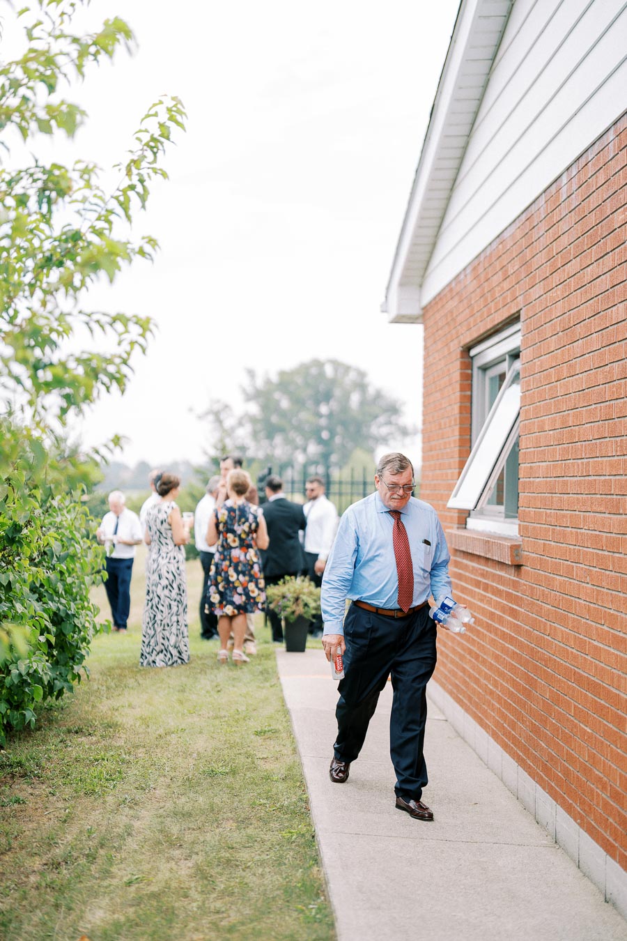 Senior man walking beside a brick building holding drinks, with a group of people socializing in the background at an outdoor gathering.