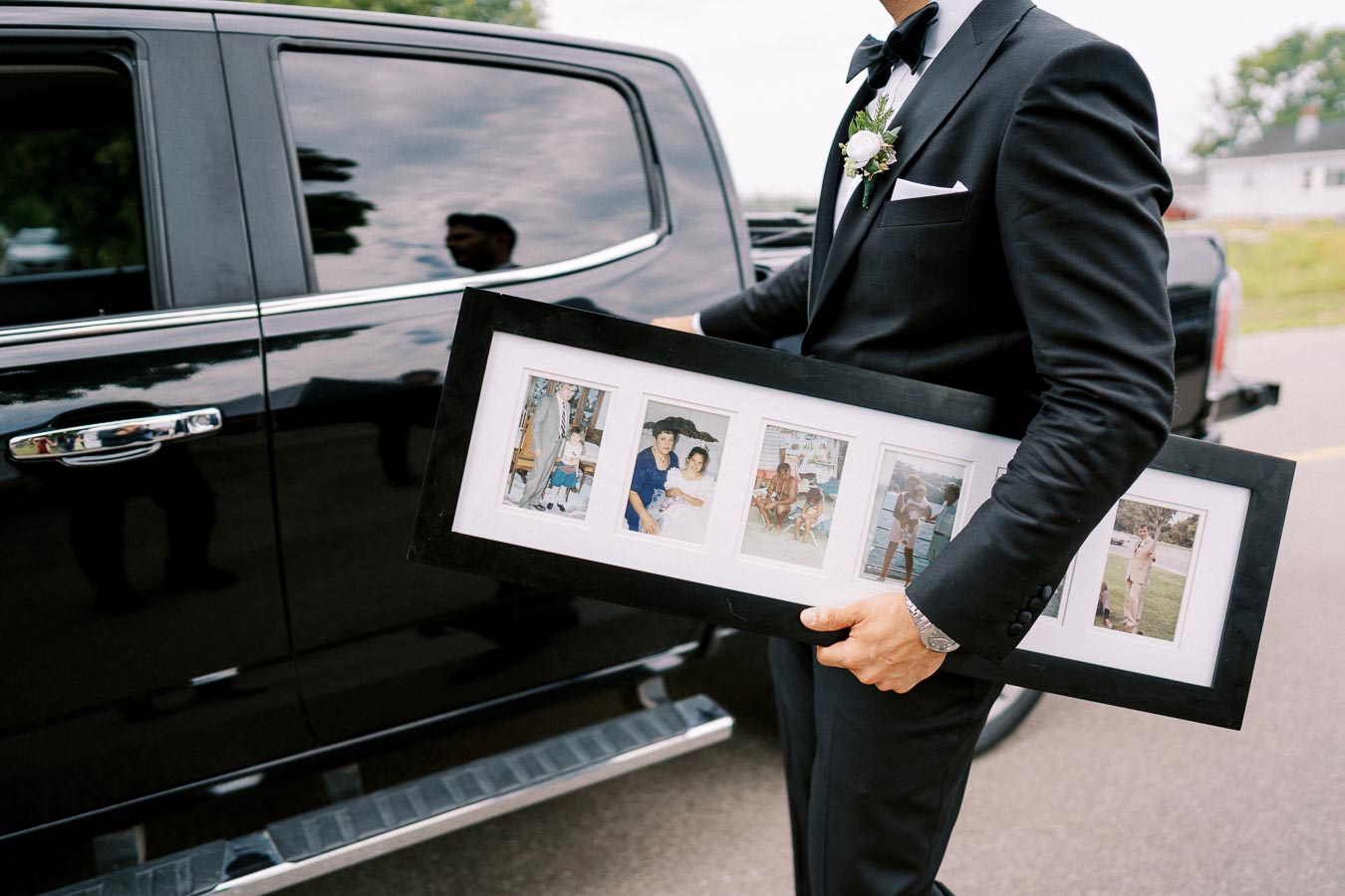 A man in a black suit holding a large framed collage of family photos beside a shiny black car, capturing a formal event or celebration.