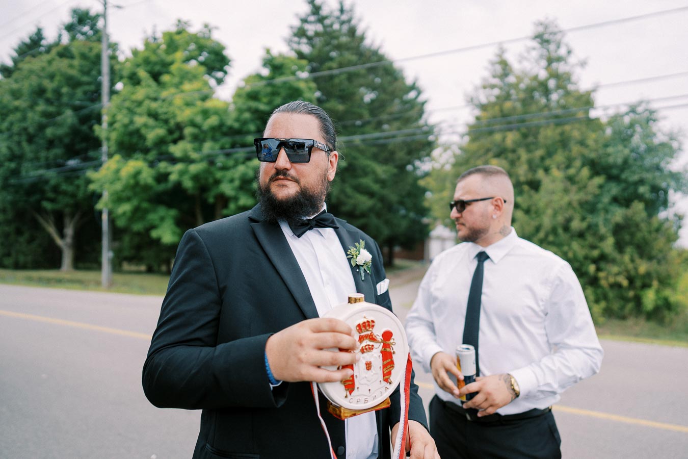Two men dressed in formal attire stand outdoors by a road, one holding a decorative flask. They're wearing sunglasses, and trees line the background, suggesting a celebratory or wedding event.