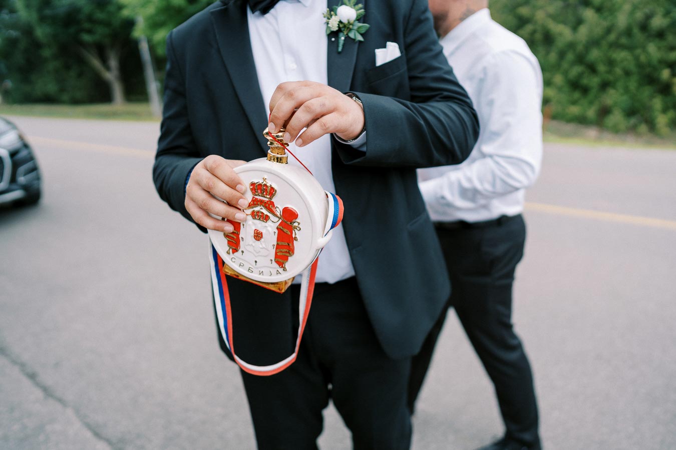 A man in a suit holding a ceremonial flask with a Serbian emblem and tricolor ribbon, outdoors at a formal event.