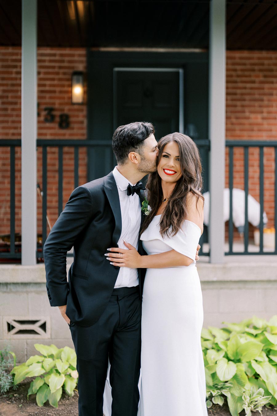 A couple dressed elegantly, with the man in a black tuxedo and the woman in a white off-shoulder dress, posing affectionately in front of a brick house with greenery.