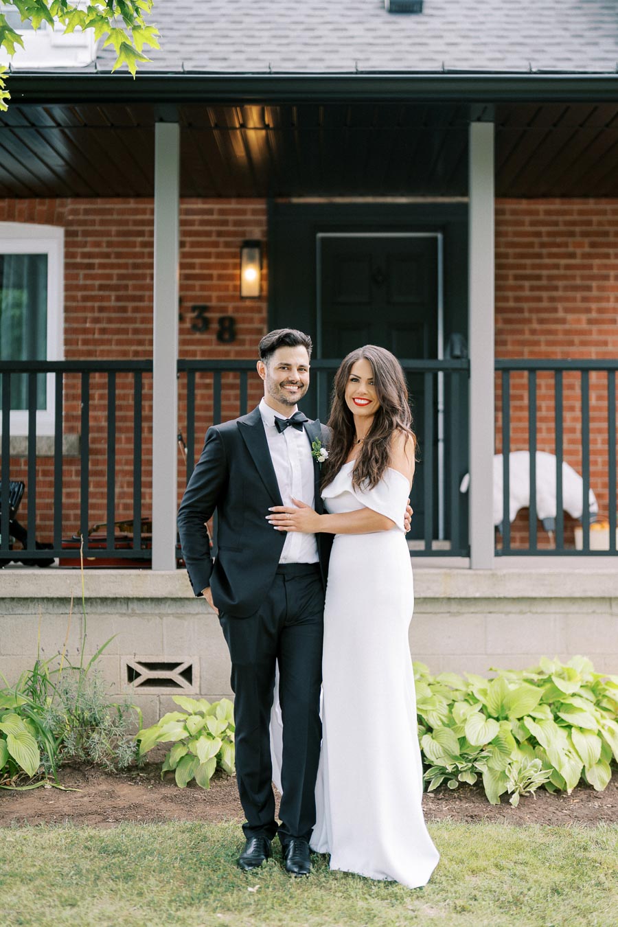 Elegant couple in formal attire posing happily outside a brick house, surrounded by greenery, showcasing a joyful moment.