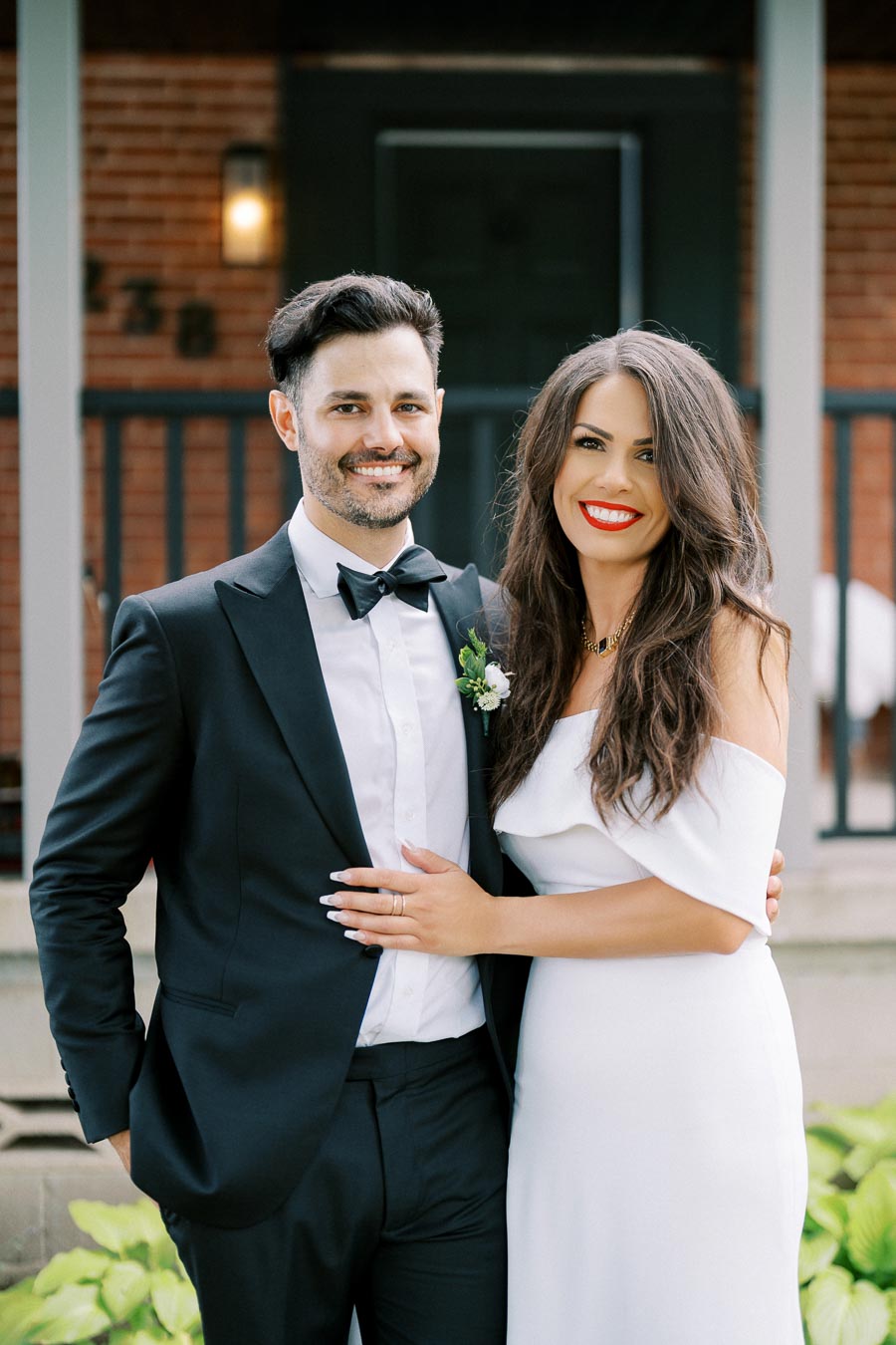 Elegant couple smiling in formal attire, with the man in a classic black tuxedo and the woman in a stylish white dress, standing in front of a brick building.