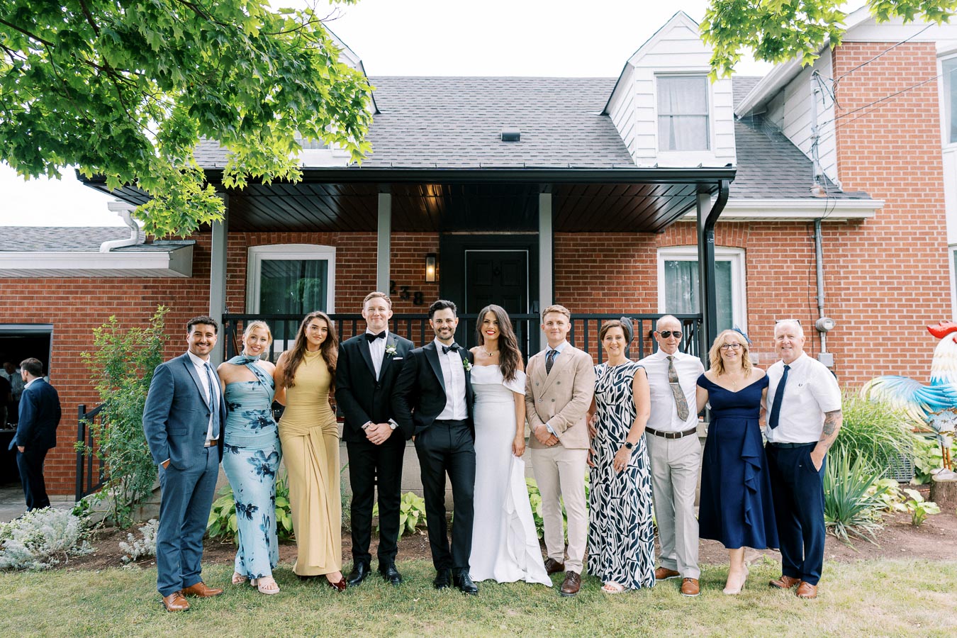 A group of well-dressed people, including a bride and groom, standing together outside a brick house, celebrating a special occasion such as a wedding, with greenery and laughter all around.