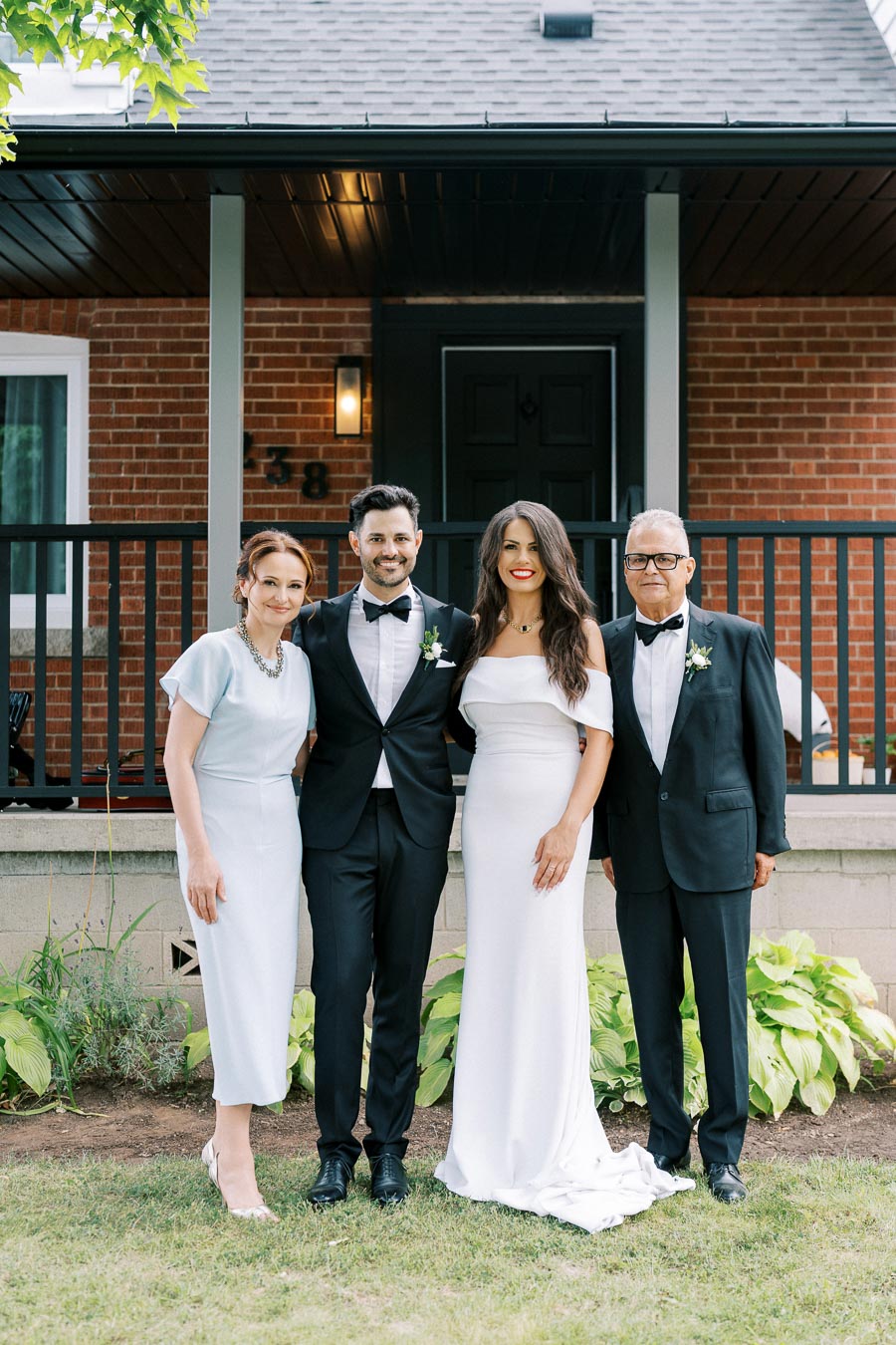 Elegant wedding party posing in formal attire in front of a brick house, featuring two women in white dresses and two men in black tuxedos with bow ties, standing on a lush green lawn.