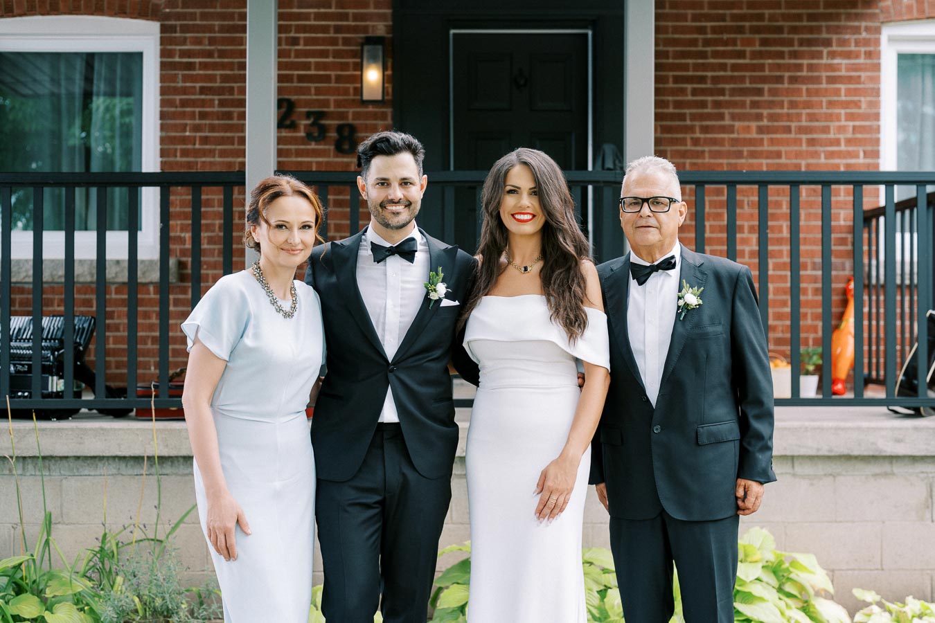 A group of four people dressed formally, smiling in front of a brick house. Two men in tuxedos stand beside two women in elegant dresses.