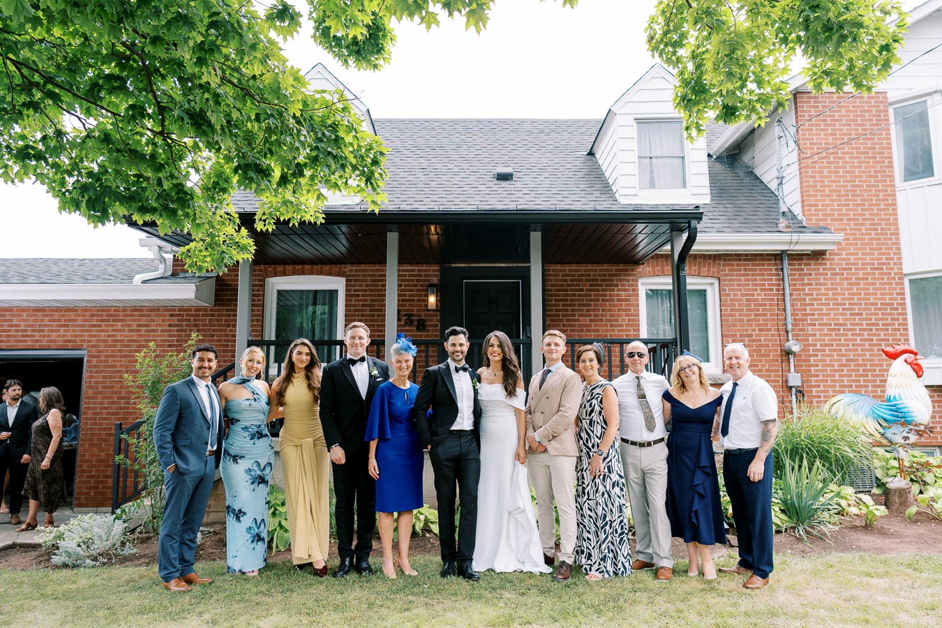 Group of elegantly dressed people posing together outside a red brick house, with trees providing shade.