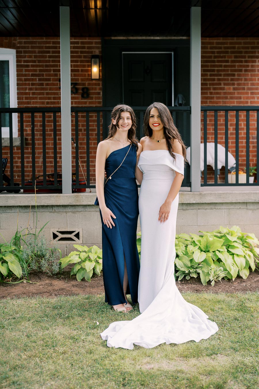 Two women elegantly dressed in formal gowns, one in navy blue and the other in white, standing in front of a brick house with a porch, surrounded by greenery.