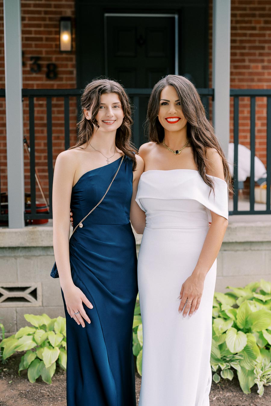 Two women dressed elegantly, one in a navy blue gown and the other in a white off-shoulder dress, smiling in front of a red brick house with a black door and green plants in the foreground.