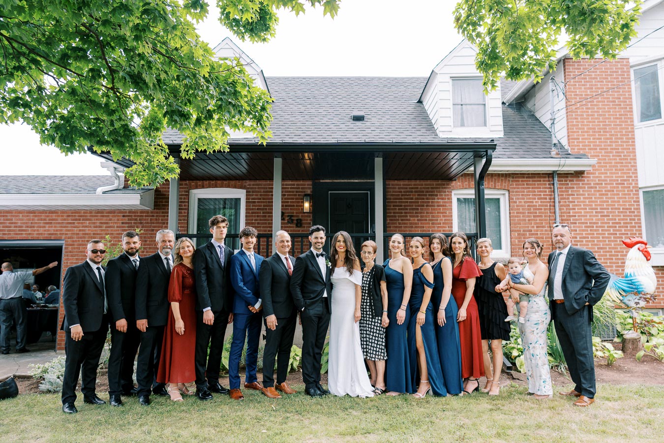 Group of well-dressed people, including a bride in a white dress, posing together in front of a red brick house under a tree.
