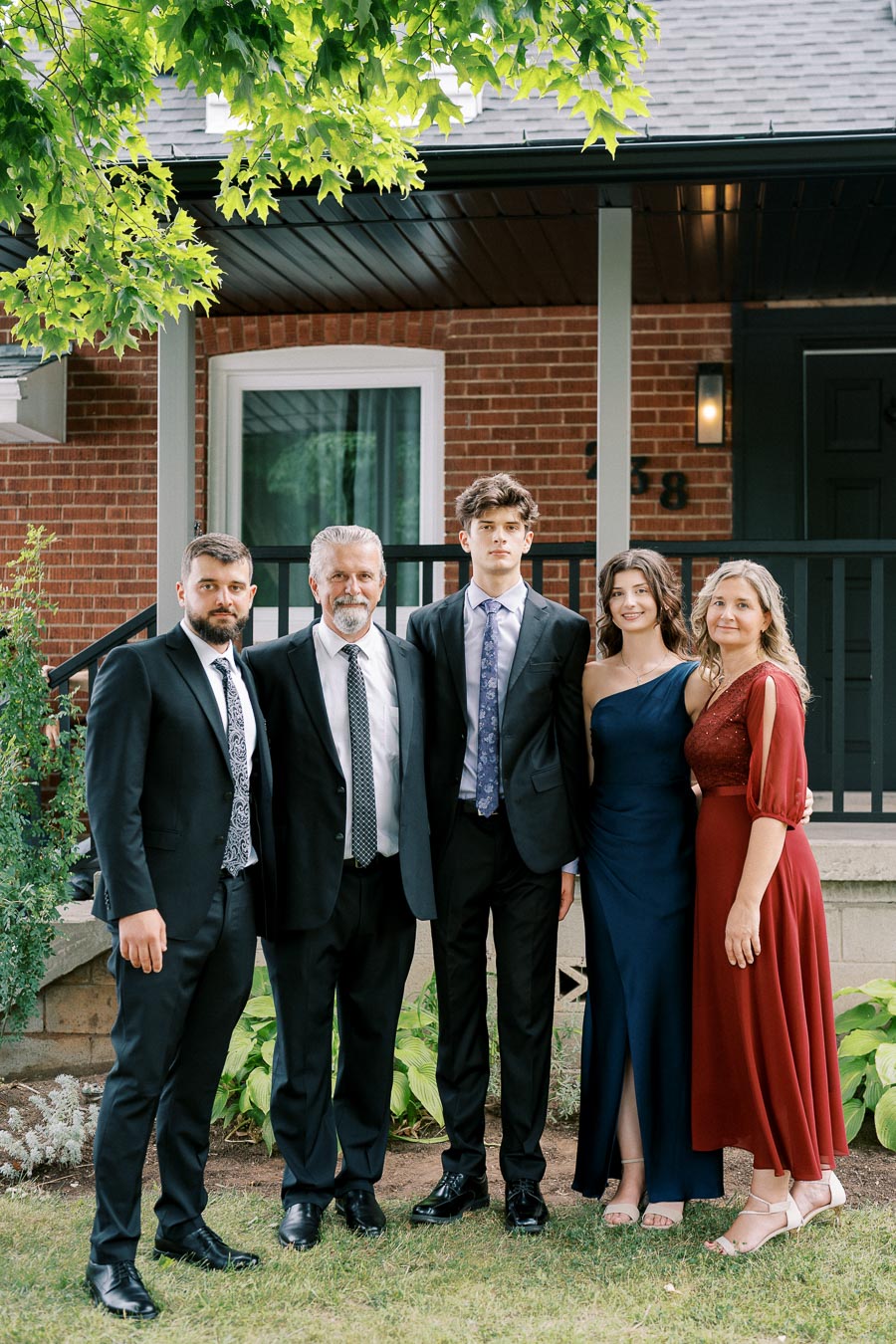 A family dressed in formal attire poses together outside a brick house, with lush greenery in the background.