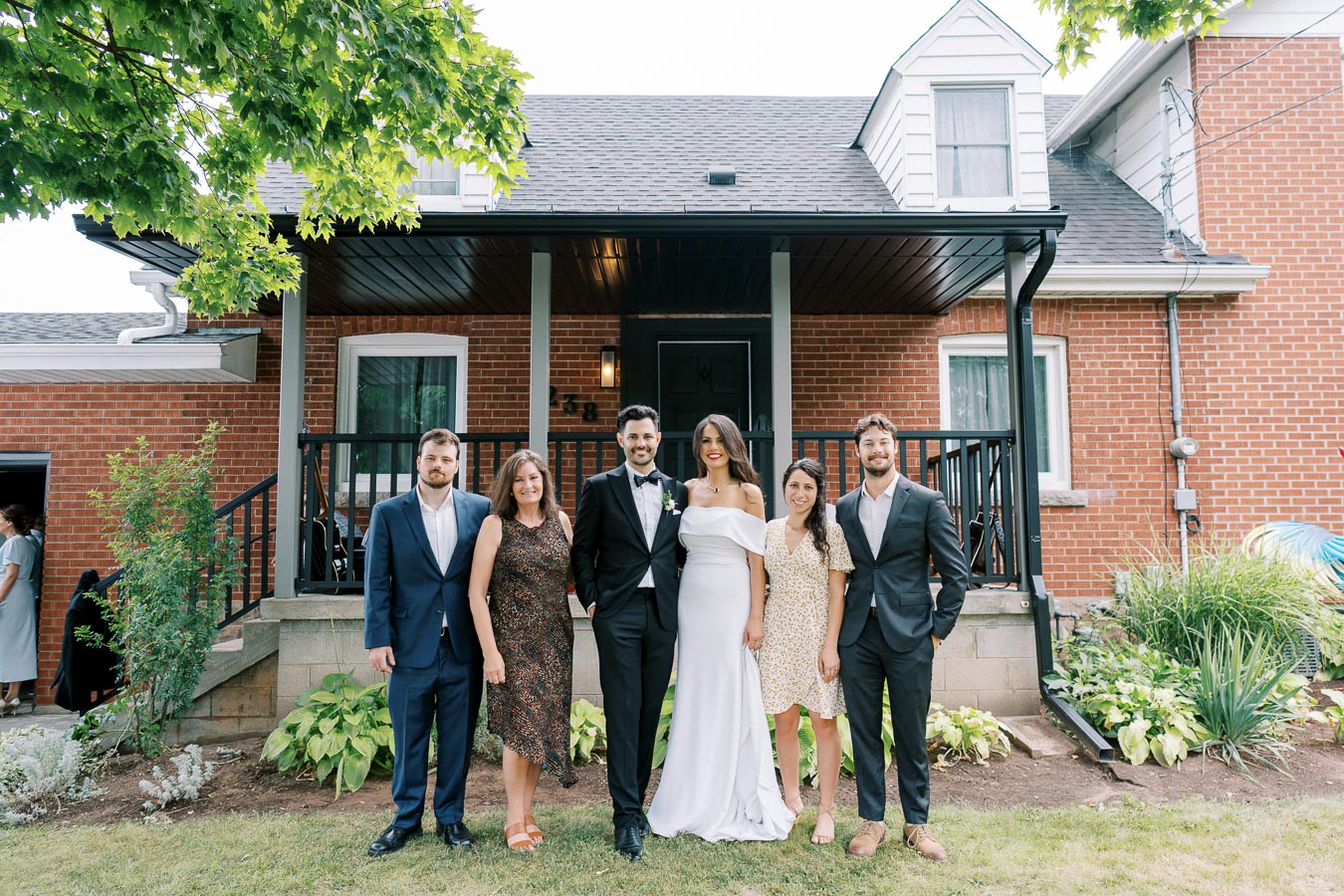 A group of people in formal attire posing outside a red brick house with a porch. The women are wearing dresses, one in a white gown, while the men are in suits. The setting features greenery and a sunny day. Perfect for wedding or family gathering themes.