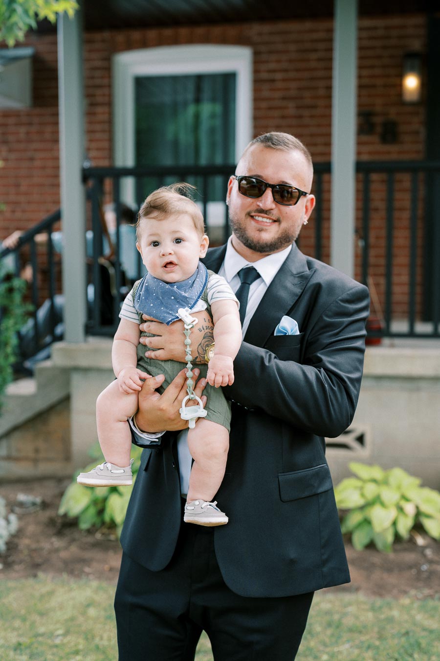 A man in a suit and sunglasses holding a baby dressed in casual attire with a pacifier clip, standing in front of a brick house with a porch.