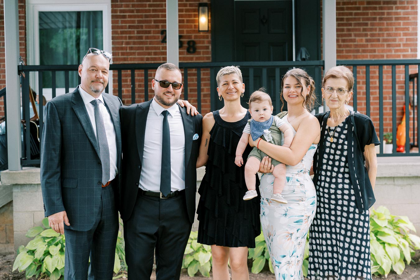 A family of six, including two men in suits, two women in dresses, one holding a baby, and an elderly woman, smiling in front of a brick house with a porch.