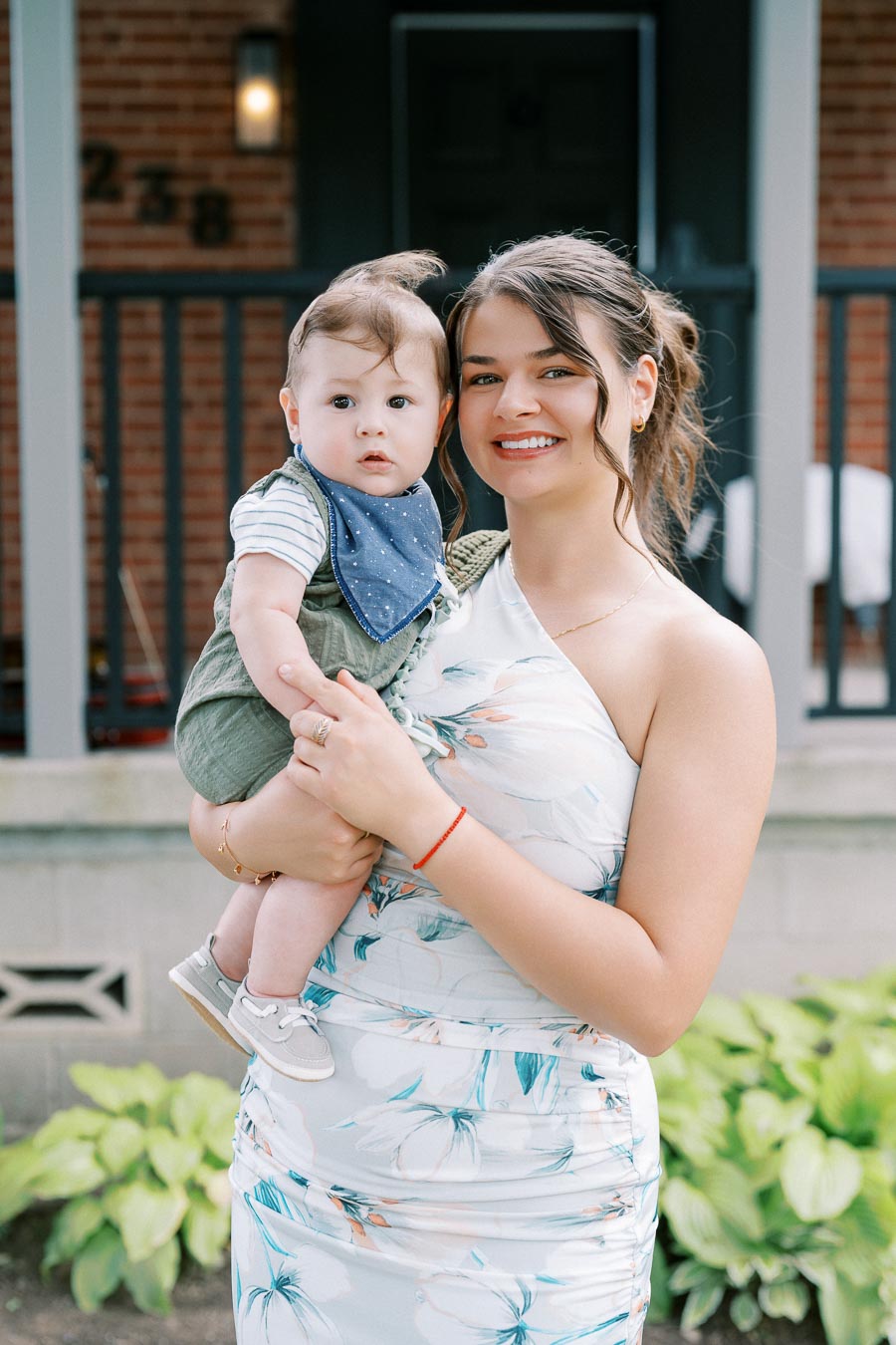 Smiling young woman holding a baby in front of a porch, both dressed in summer attire, with greenery in the background.
