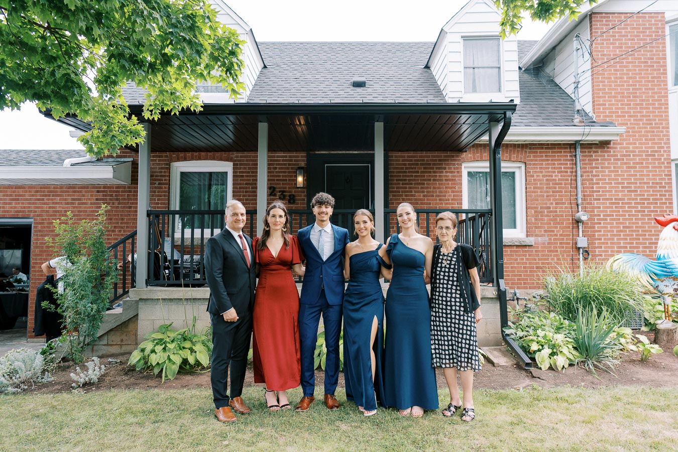 A group of six people in formal attire pose in front of a brick house with a porch, surrounded by green plants and landscaping, under a clear sky.