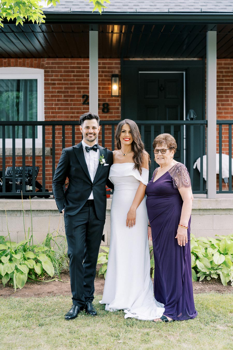 A groom in a black tuxedo, a bride in a white gown, and an older woman in a purple dress pose smiling in front of a brick house with black railings and greenery around them.