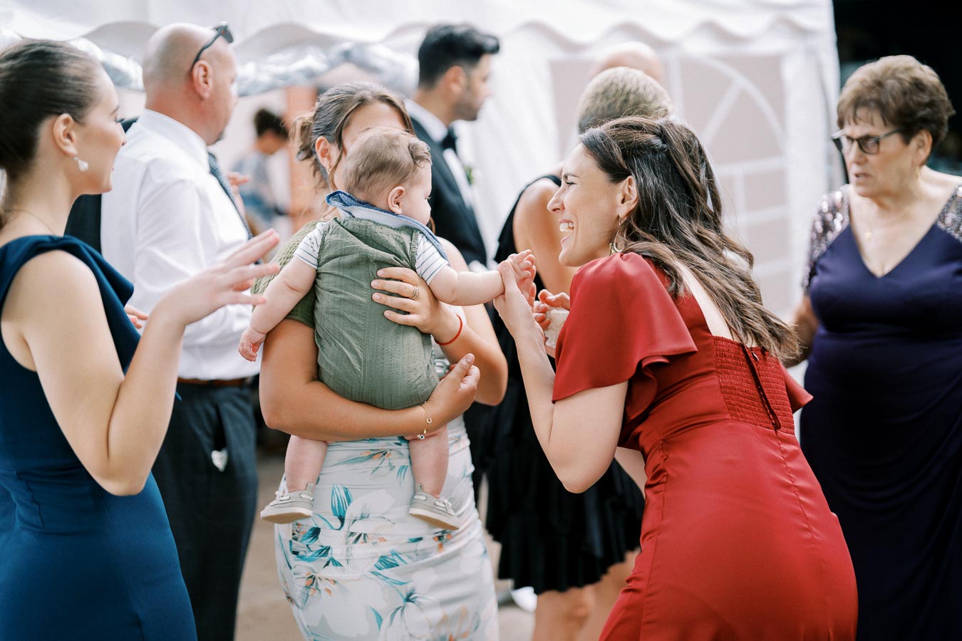 A joyous gathering at a celebration, featuring a woman in a red dress interacting warmly with a baby held by another guest, surrounded by people in elegant attire.