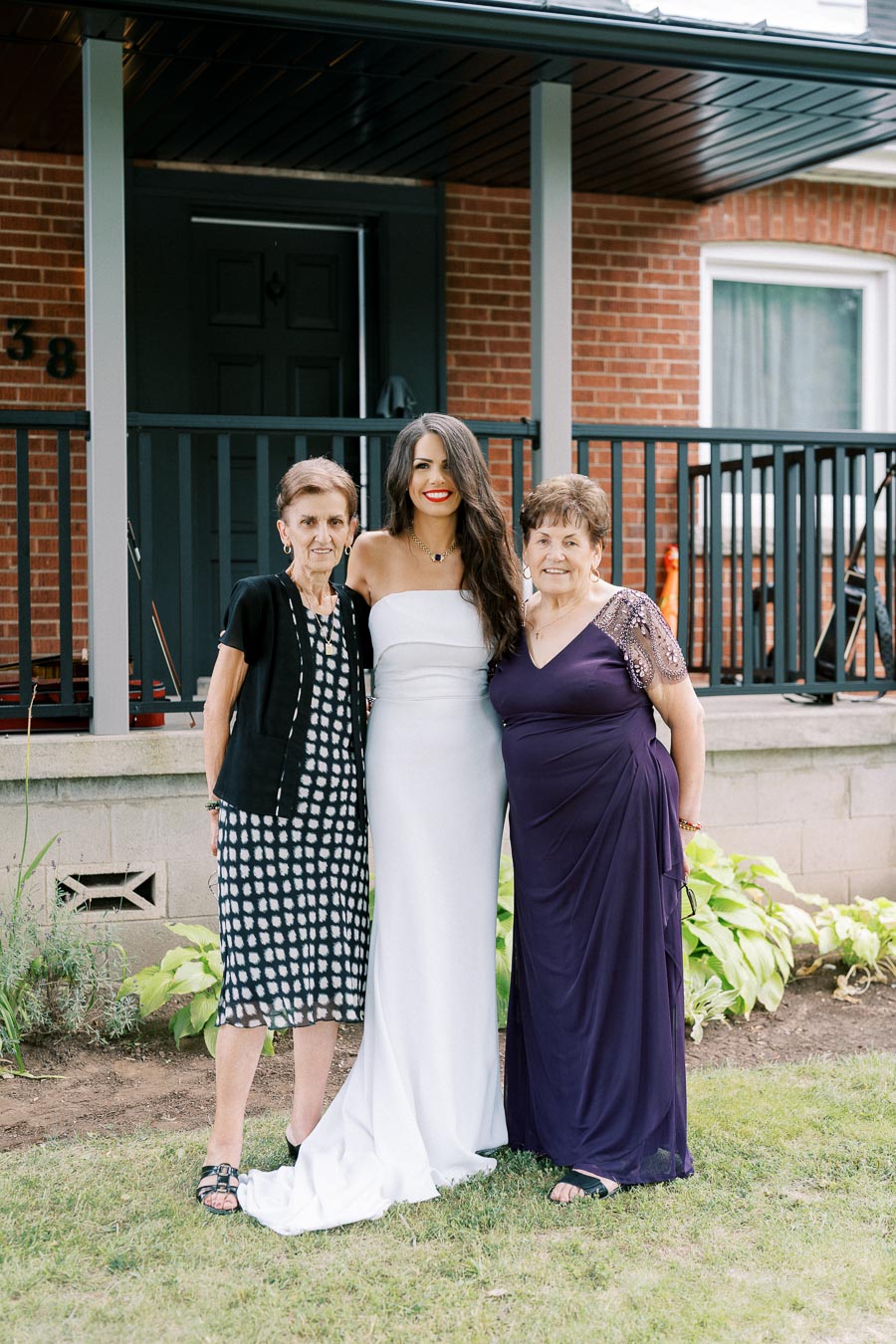 Three women standing together smiling in front of a brick house, with the middle woman wearing a white dress and the other two in patterned and purple dresses.
