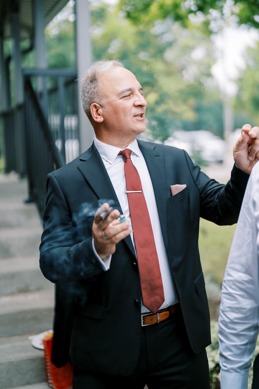 A well-dressed man in a suit with a red tie holding a smoking cigar outside on a staircase, surrounded by greenery.