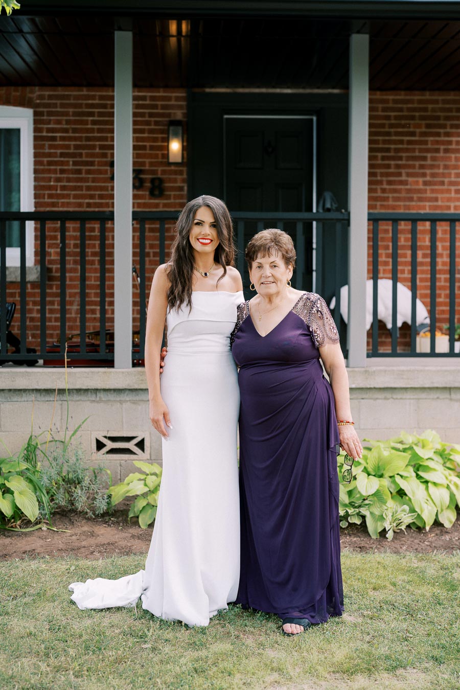 Two women standing together outside a brick house, one wearing a white dress and the other in a purple gown. They are smiling and posing in front of a porch.