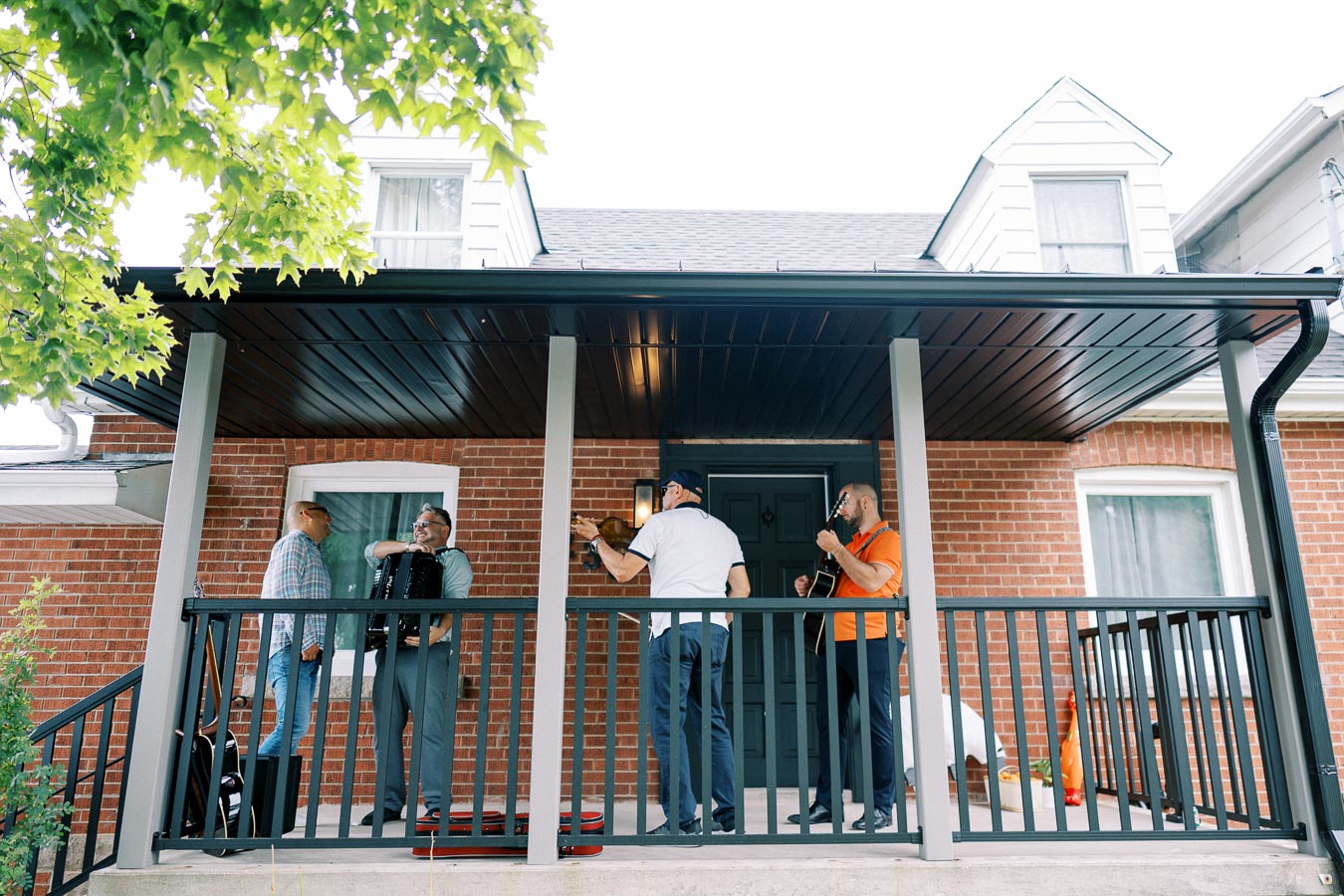 Four men playing musical instruments on a house porch, featuring a violin, an accordion, and a guitar, with brick walls and a tree in the foreground.