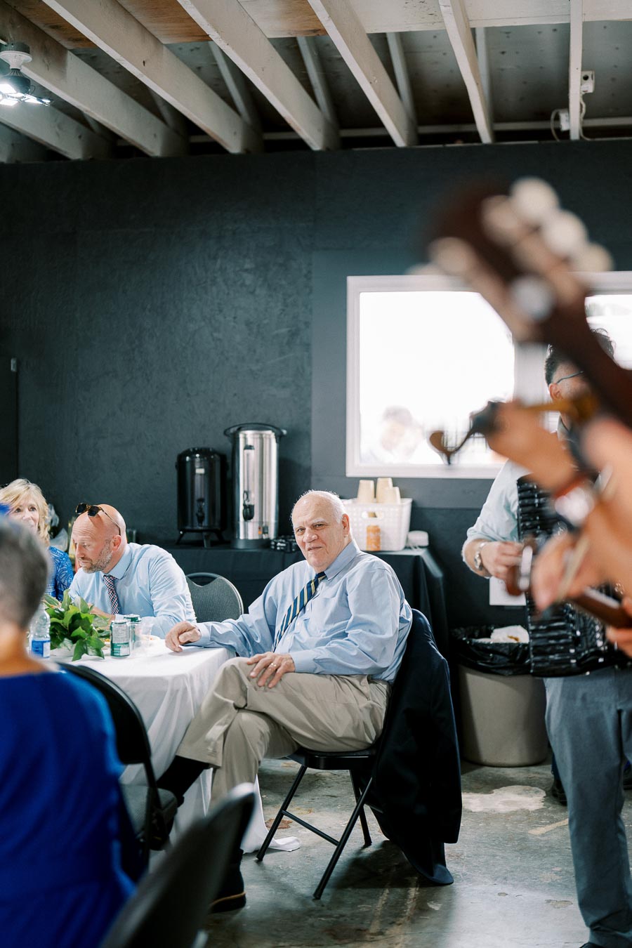 People gathered at an indoor event, sitting at a table with a white tablecloth, listening to a musician playing a guitar. Beverage dispensers and cups are in the background, indicating a casual gathering or celebration.