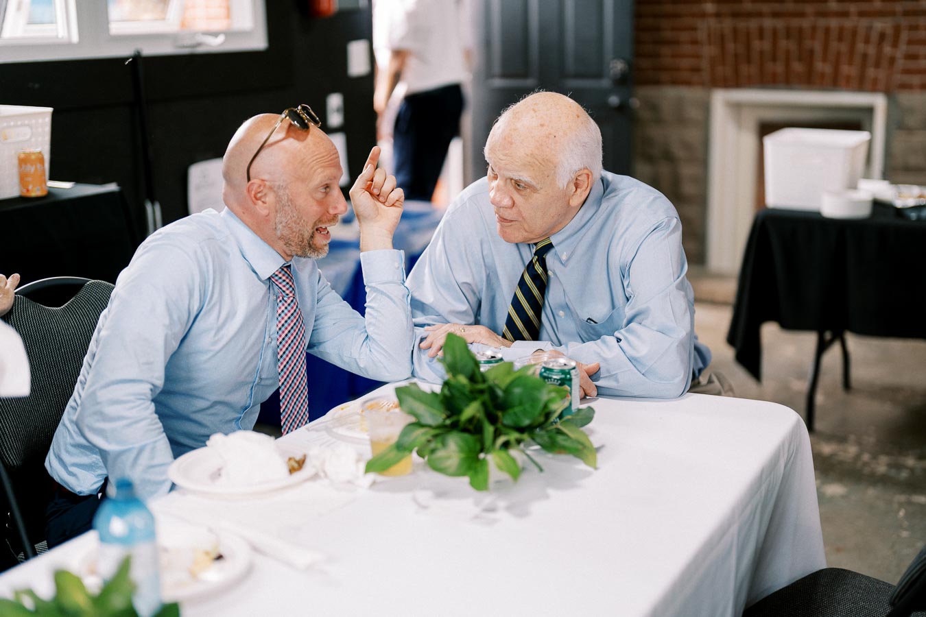 Two men in conversation at a dining table, both wearing light blue shirts and seated indoors. On the table are plates, a potted plant, and beverages, suggesting a casual meeting or gathering environment.
