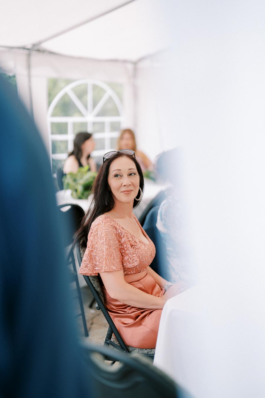 A woman in a coral dress smiling while seated at an outdoor event with a tent canopy overhead, surrounded by guests and white tables.