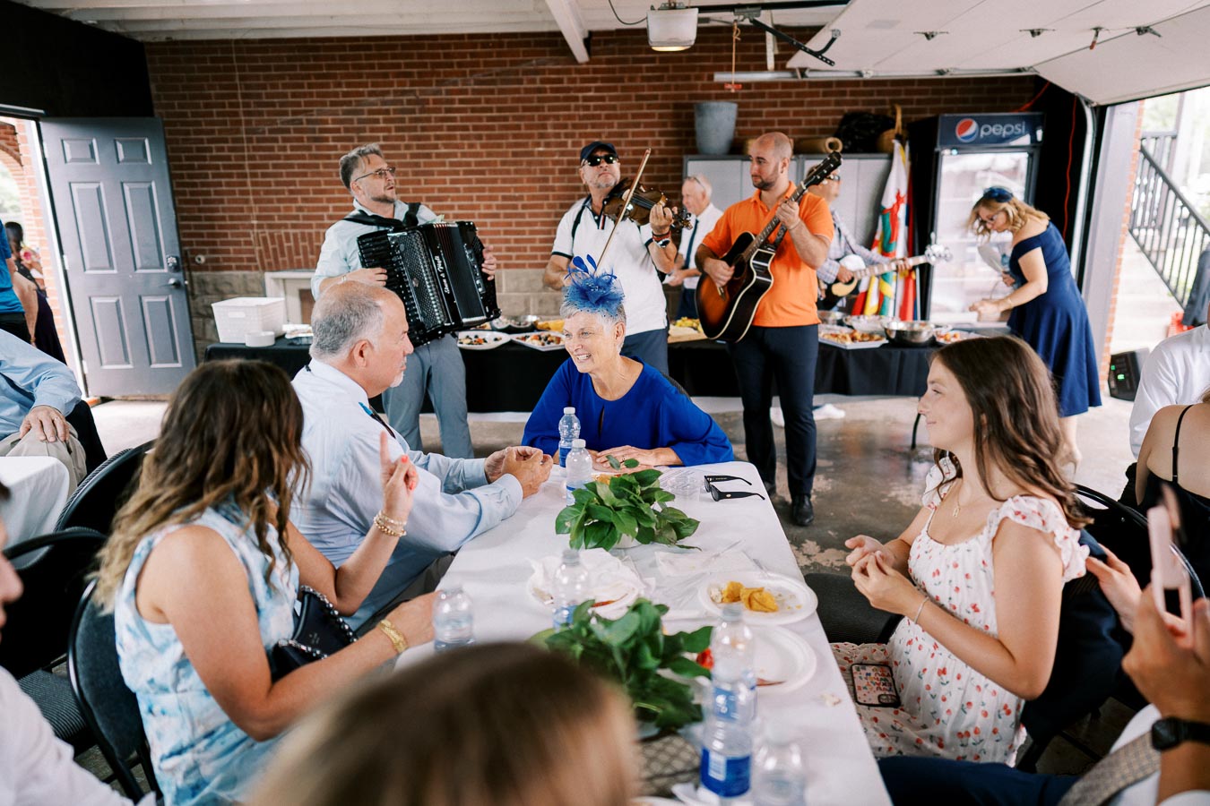 A lively indoor gathering with a group of people sitting around a table enjoying a meal. A live band featuring accordion, violin, and guitar players performs in the background, adding to the festive atmosphere. The setting includes brick walls and a buffet table with food.