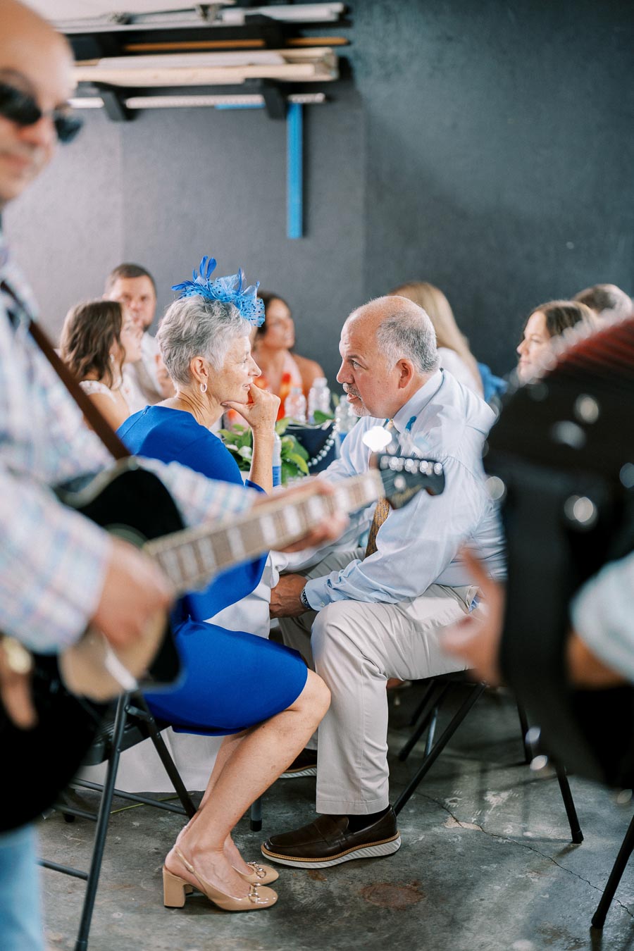 Elderly couple in formal attire conversing at a social gathering, with a musician playing guitar in the foreground.
