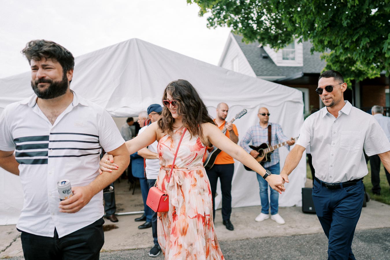 A group of people enjoying an outdoor celebration near a white tent, with music playing and a woman in a floral dress holding hands with two men.