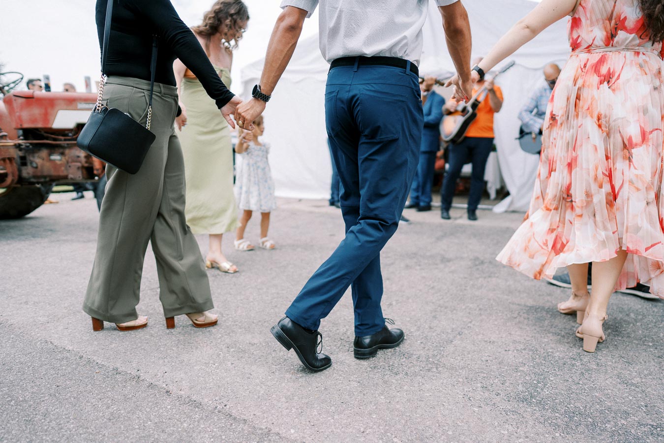 A group of people holding hands in a circle dancing outdoors, wearing colorful clothing, with a band playing in the background.