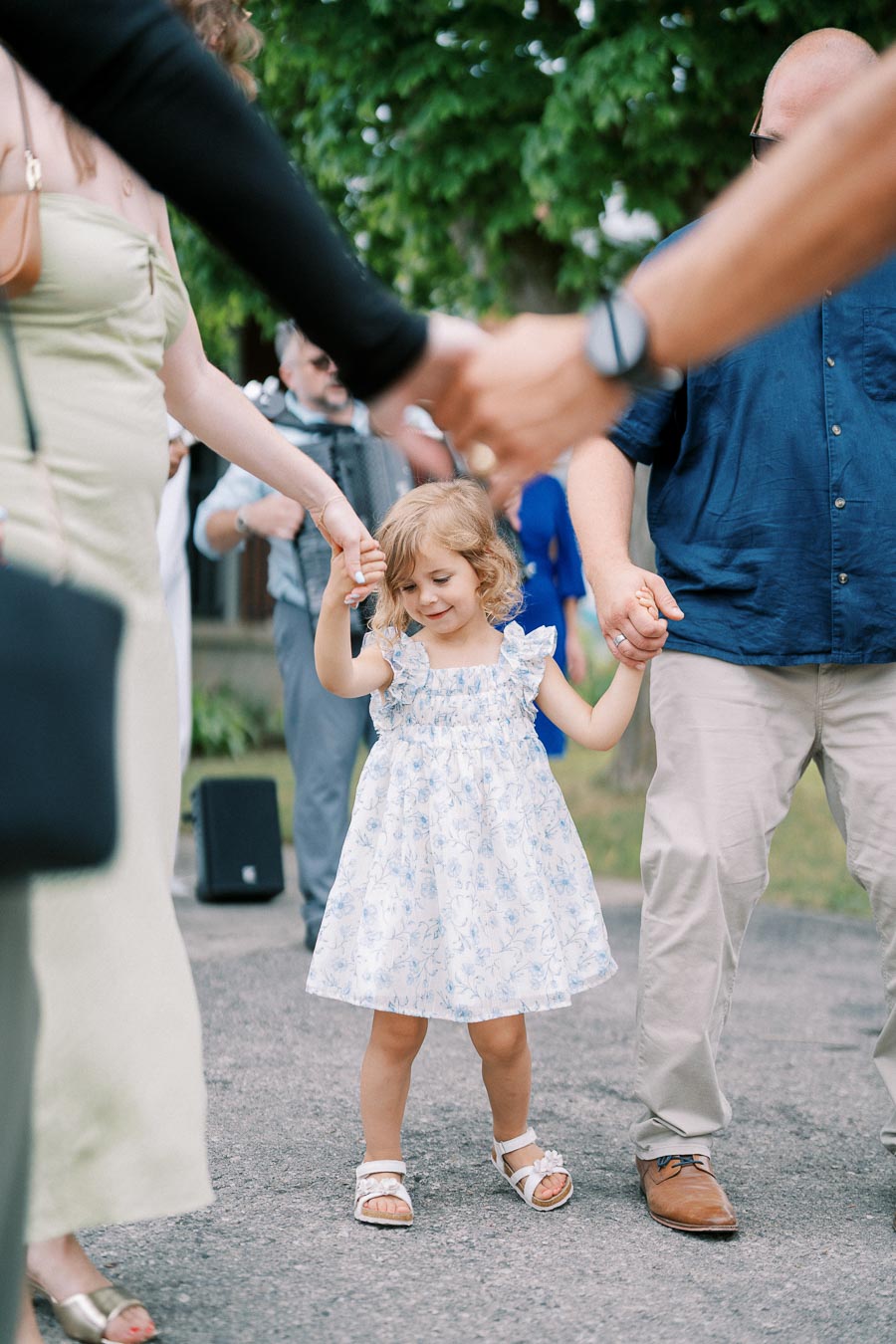 A small child in a floral dress joyfully holds hands with adults, participating in a lively outdoor dance circle at a summer gathering, with greenery in the background.