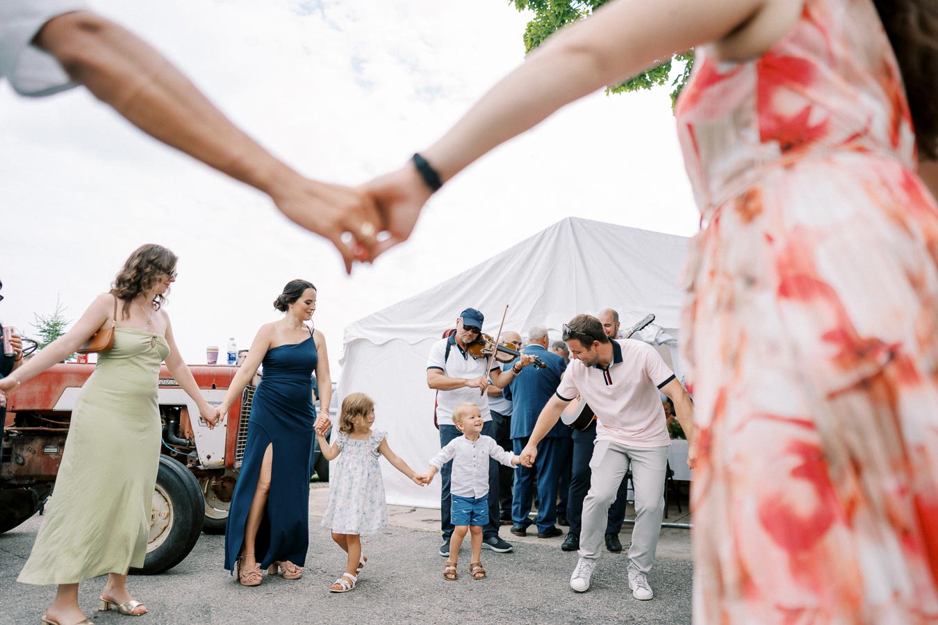 People joyfully dancing in a circle outdoors, holding hands with children, with musicians playing instruments in the background, under a tent on a sunny day.