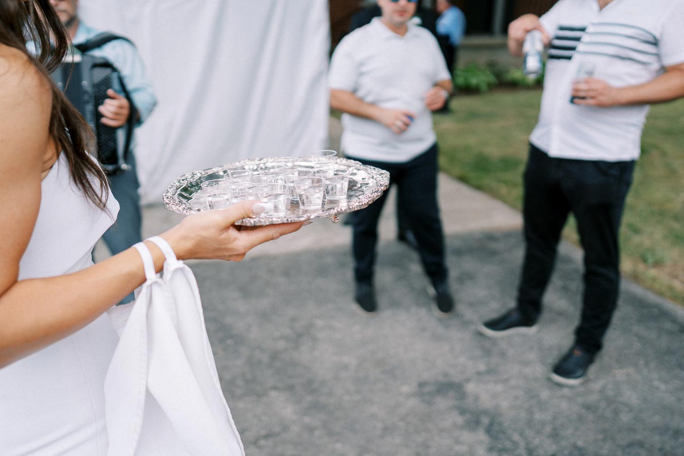 A woman in a white dress holding a silver tray with multiple shot glasses outdoors, with three men in white shirts and dark pants in the background.