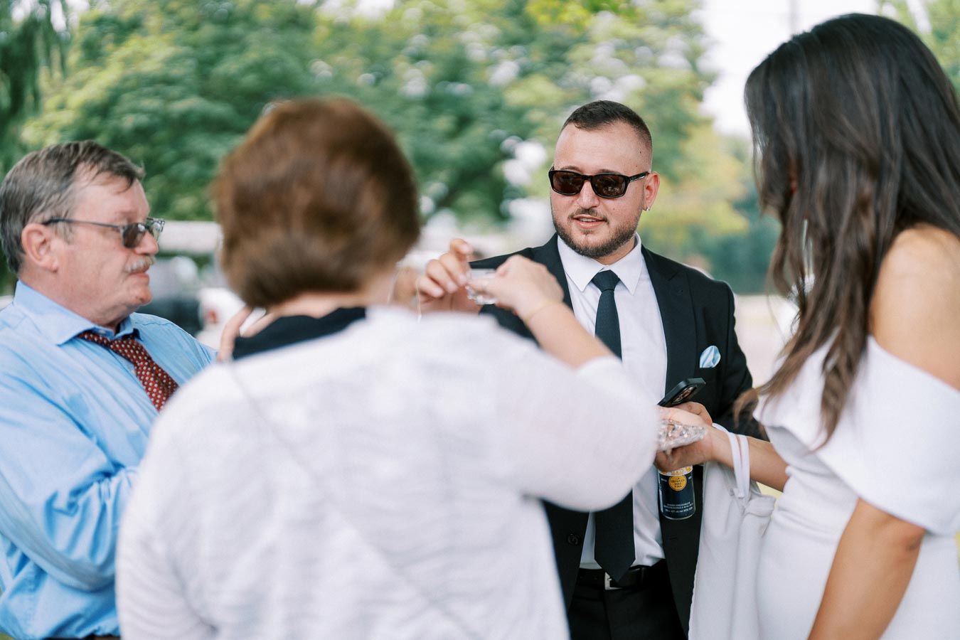 A group of people interacting at an outdoor event, with a man in a suit and sunglasses engaging in conversation, surrounded by others in formal attire.