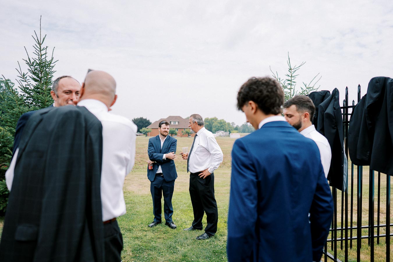 Group of men in suits talking outdoors at a formal event, with jackets hanging on a fence nearby.