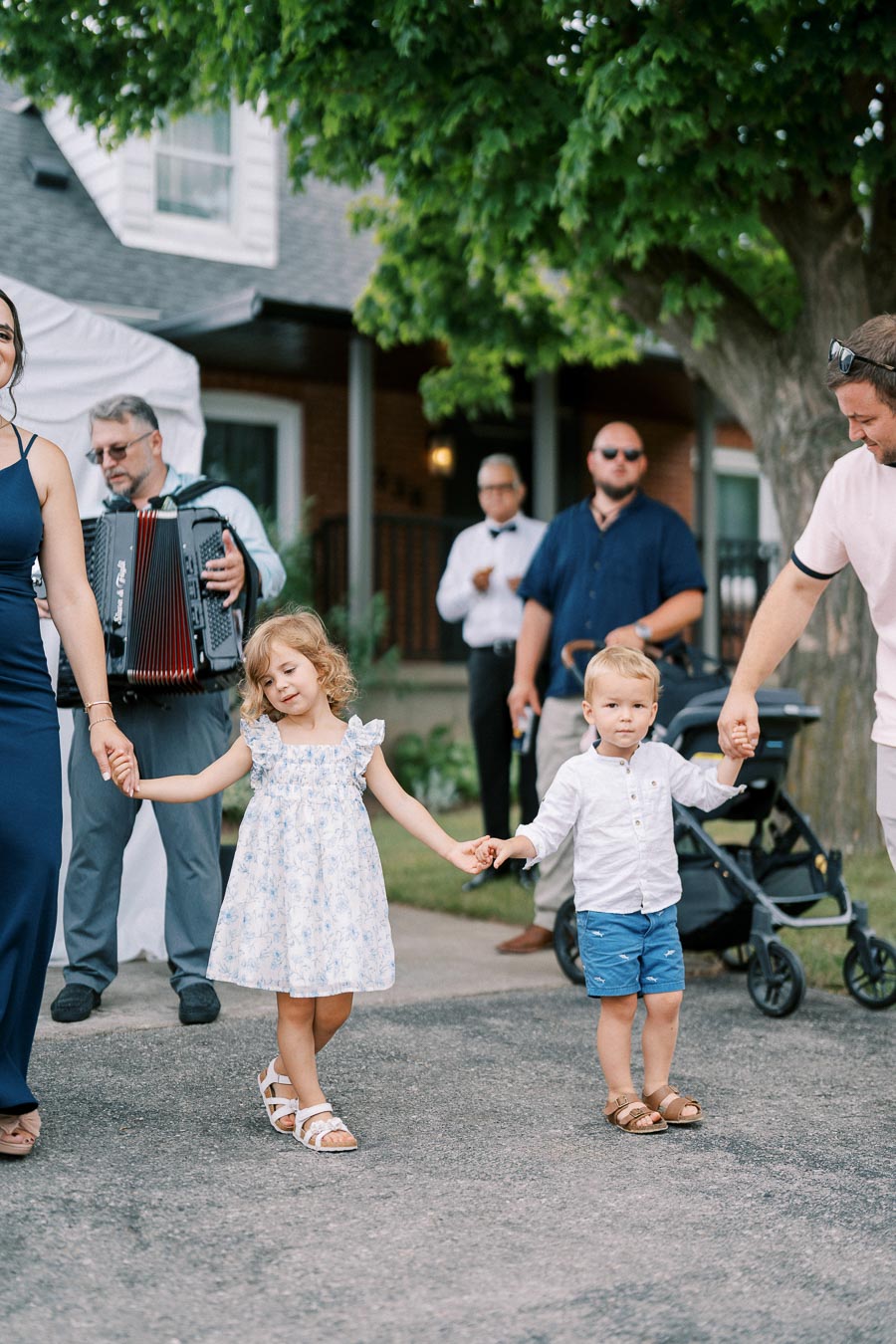 Children holding hands at an outdoor gathering, surrounded by adults and musicians, with a tree and stroller in the background.