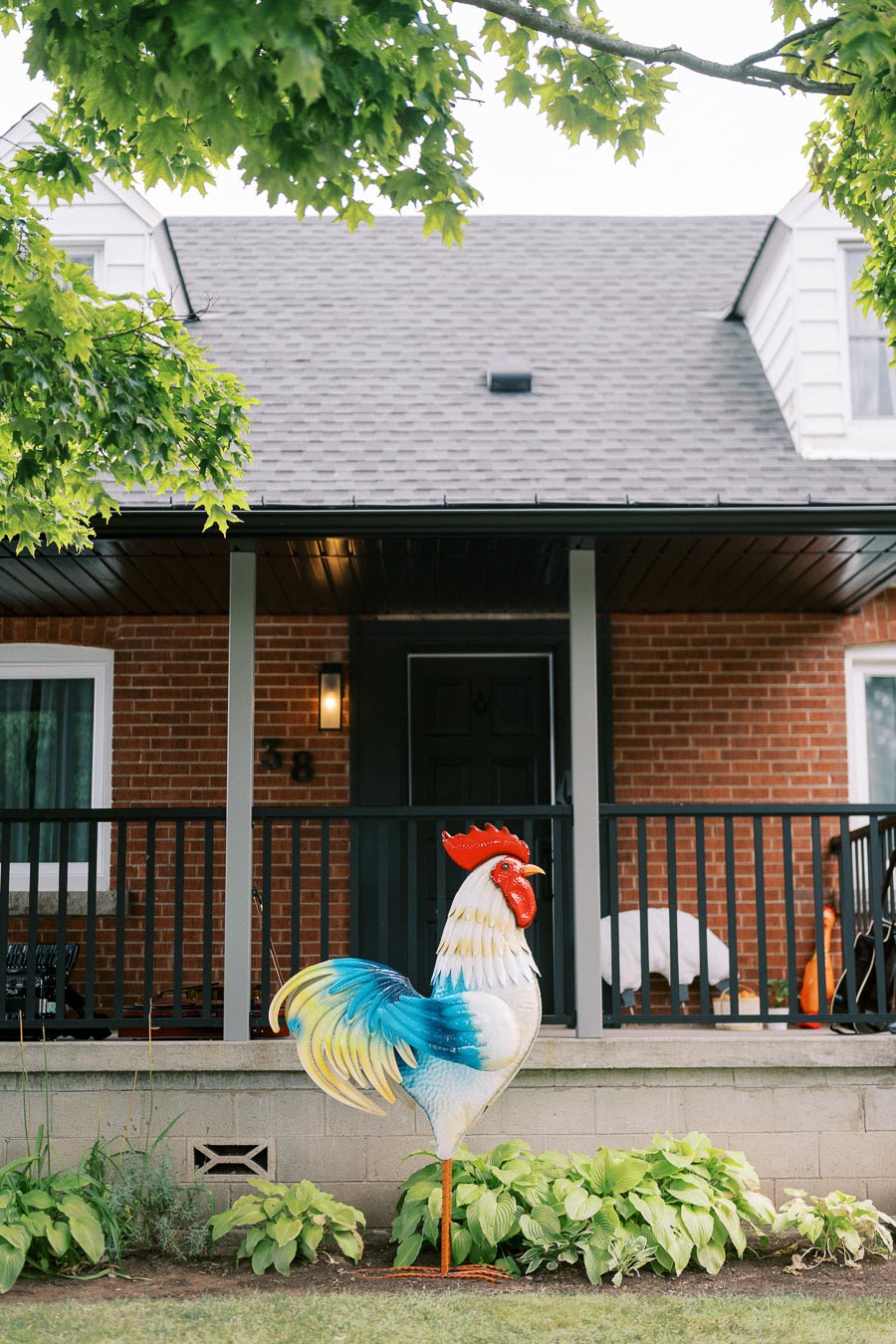 Colorful metal rooster sculpture in front of a red brick house with a gray roof and black railing porch, surrounded by lush green plants and vibrant leafy trees.