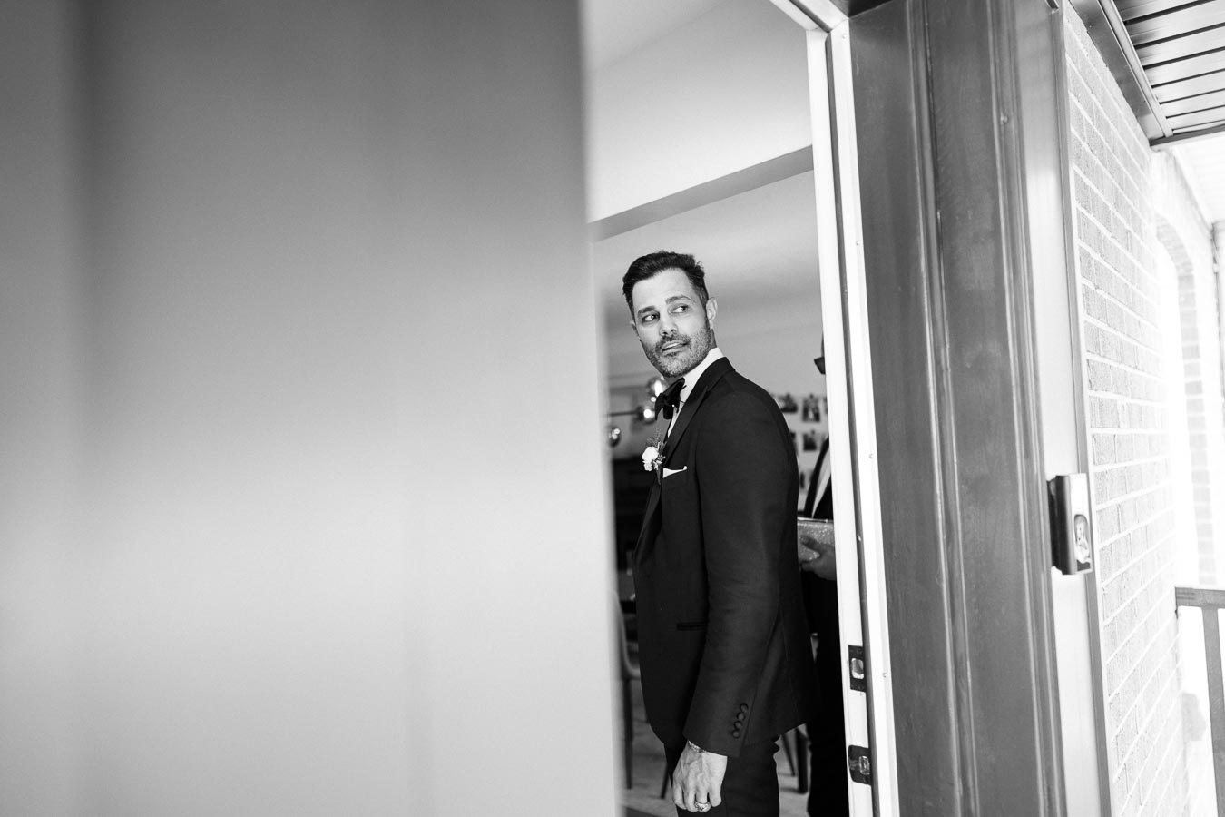 Black and white photo of a man in a suit with a boutonniere, standing in a doorway and looking back, capturing an elegant and formal moment.