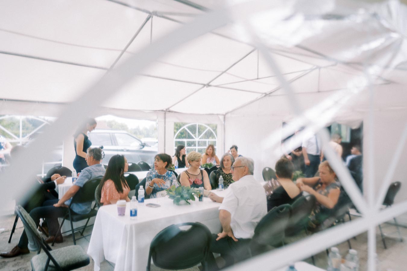 Group of people enjoying a social gathering under a white canopy tent, seated around a table with drinks and decorations.