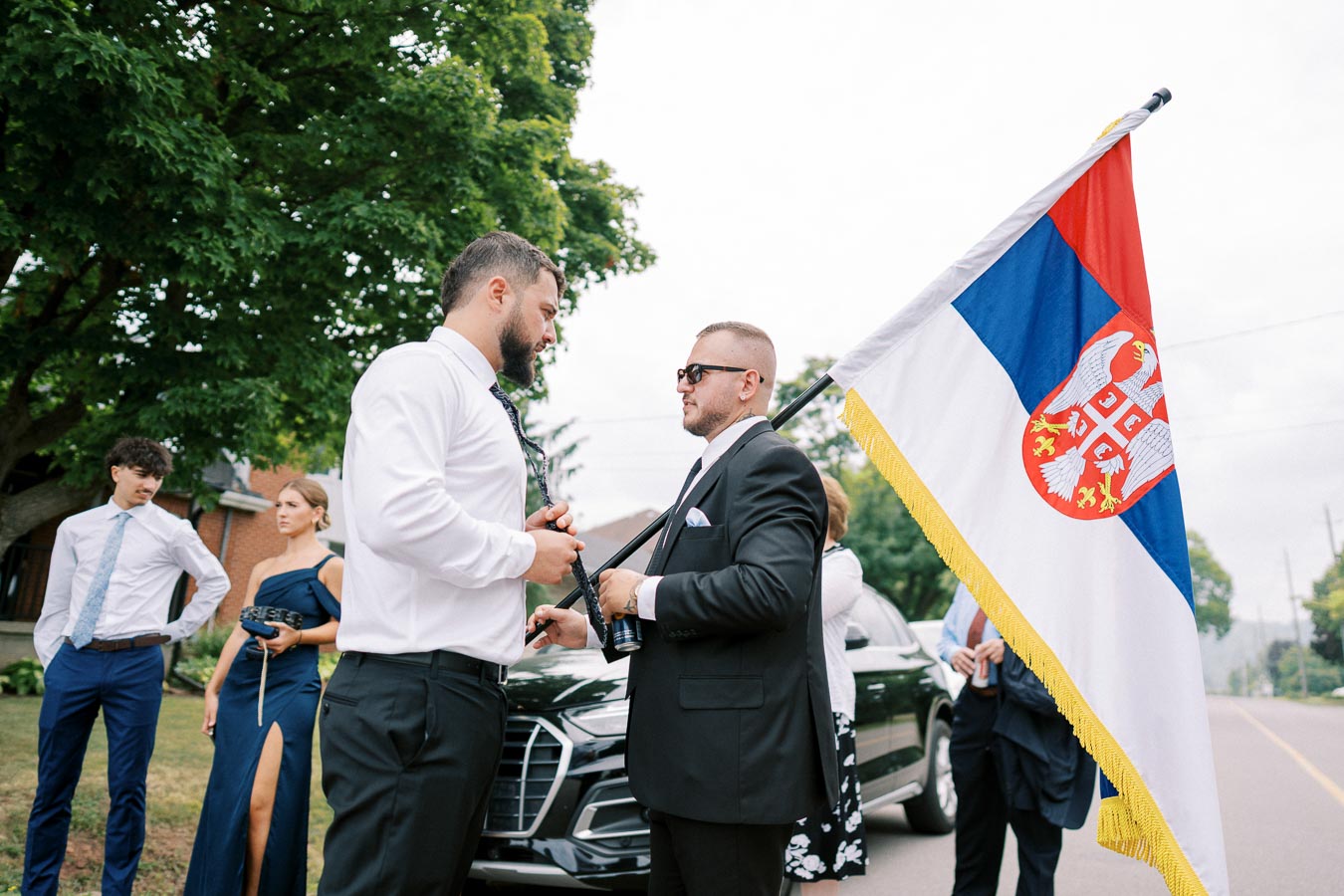 A group of well-dressed individuals standing on a street holding a Serbian flag, with cars and trees in the background.