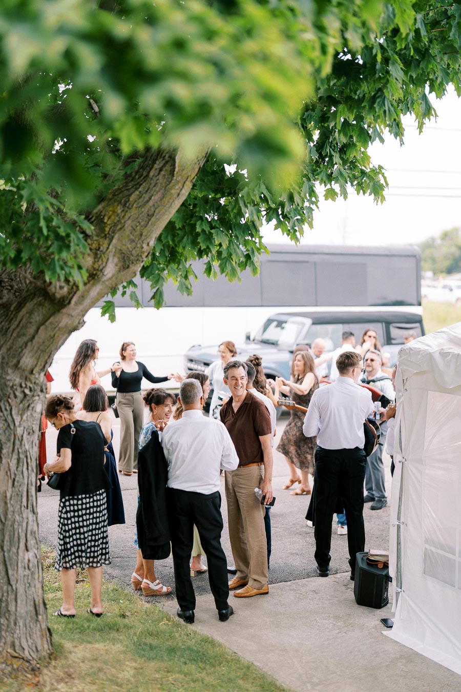 Group of people socializing outdoors under a tree at an event, with a black vehicle in the background.