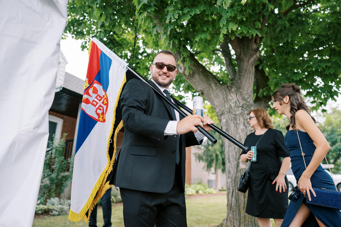 Man in a suit holding a Serbian flag at an outdoor event with women nearby.